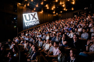 UX London conference. The room is full, and people are looking at the stage (the stage is off-screen) with apparently undivided attention. ‘UX London’ letters are projected at the far side wall in the room