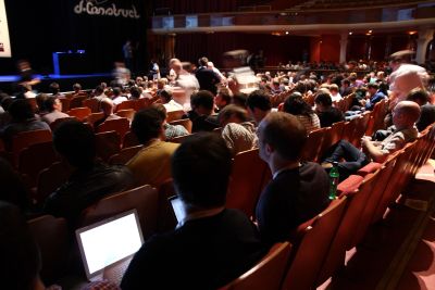The photo shows the audience at the dConstruct conference, lots of seated people in the room, some of them with their laptops open; all are looking at the stage where the ‘dConstruct’ logo is displayed