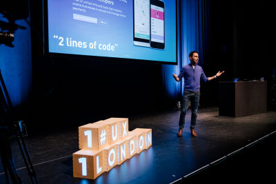 Scott Belsky speaking at the UX London conference. The ‘UX London’ logo in the form of cubes is arranged on the stage next to the speaker