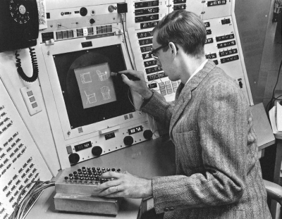 A photograph of Ivan Sutherland using SketchPad. He is drawing graphics on the screen using the light pen in his right hand while manipulating a panel of dials with his left hand. The graphic on the screen appears to be blueprints for a chair design.